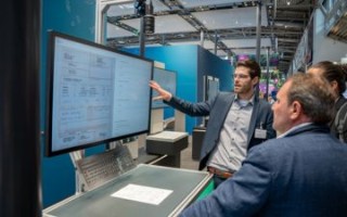 SKALA transport logisic teaser Das Bild zeigt einen Mann, der an einem Bildschirm steht und eine Präsentation oder Erklärung zu einem Thema im Bereich Transportlogistik gibt, während mehrere Zuhörer, darunter ein weiterer Mann in einem Anzug, aufmerksam zuschauen.