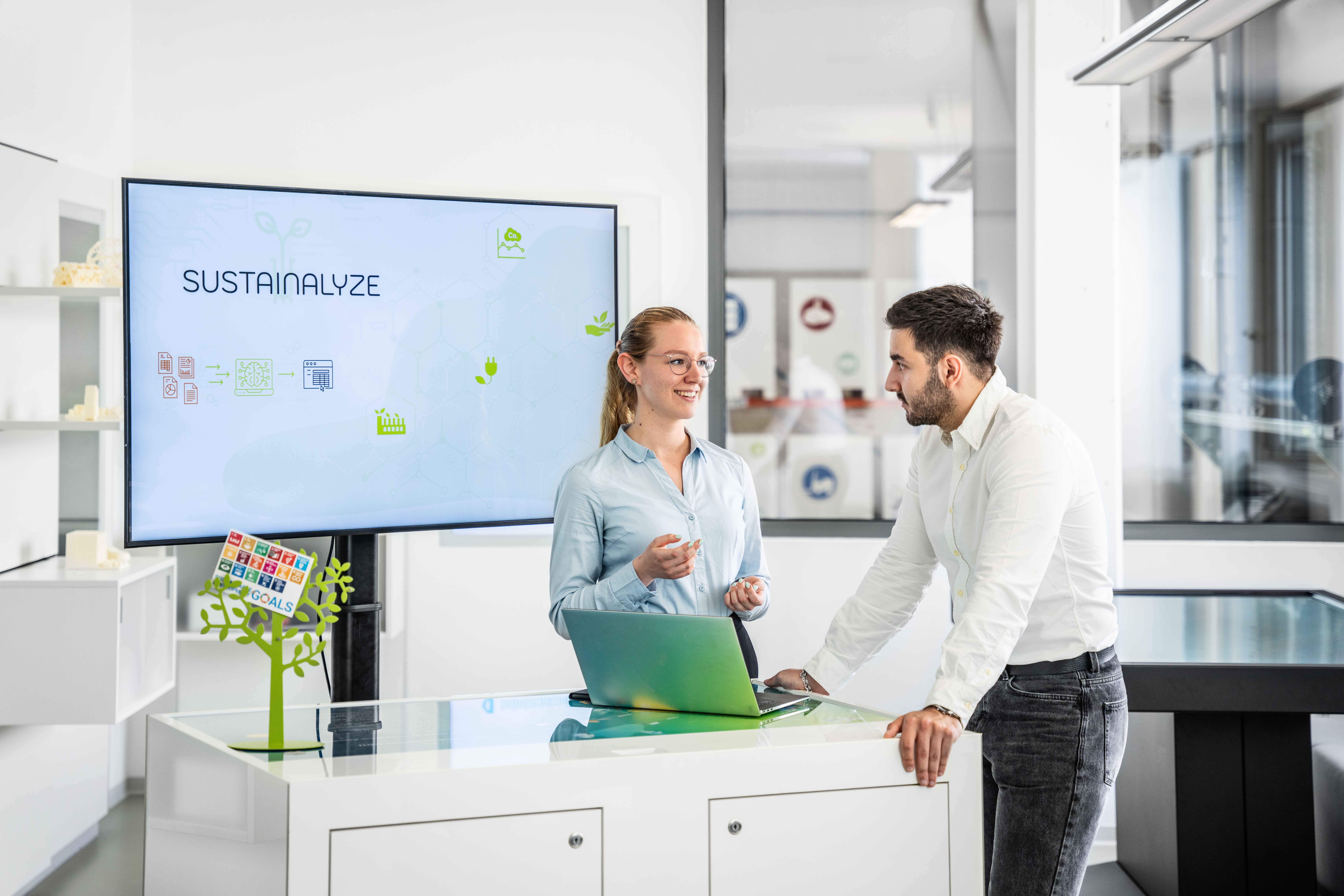 Pia Schreynemackers und Alim Coskun arbeiten am Projekt Sustainalyze, welches Nachhaltigkeitsberichte effizient bewertbar macht. das Auf dem Bild sieht man wie sie vor einer Präsentation des Projekts stehen und an einem offenen Laptop arbeiten. 