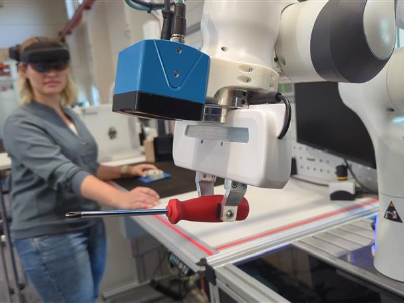 Ein Roboterarm der einen Schraubendreher hält mit einer Frau mit VR Brille im Hintergrund.