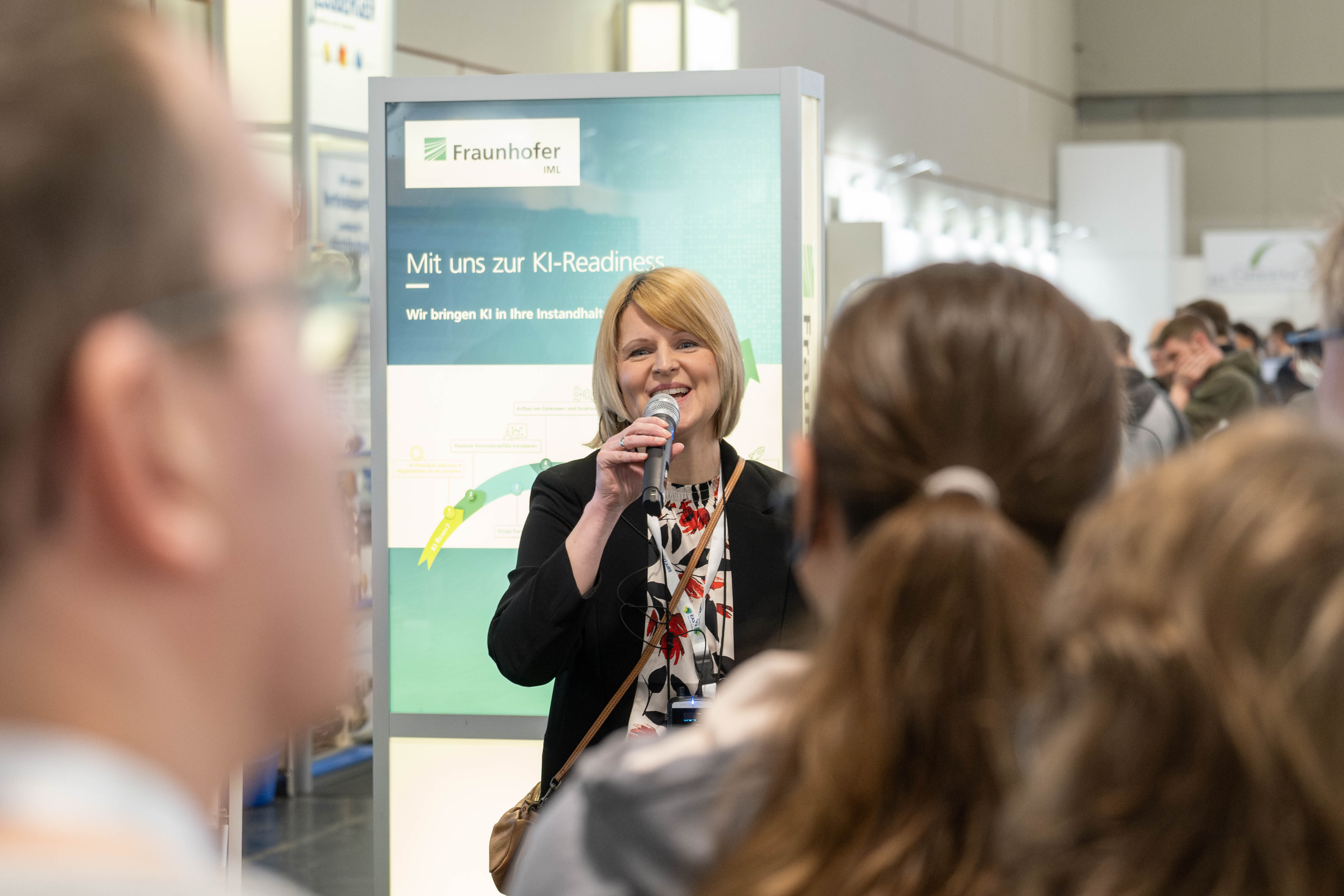 Eine Frau steht vor einem Publikum und spricht auf der maintenance Messe ins Mikrofon. Die maintenance in Dortmund ist die führende Fachveranstaltung zum Thema industrielle Instandhaltung und eine etablierte Bühne für Innovationen und Trends. Die Frau moderiert einen Messerundgang.