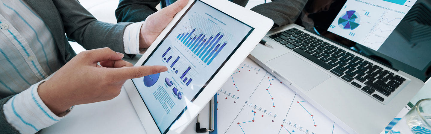 Mann im Anzug präsentiert auf einem Tablet Statistiken zum Stammdatenmanagement, während im Hintergrund ein Laptop ähnliche Daten zeigt.