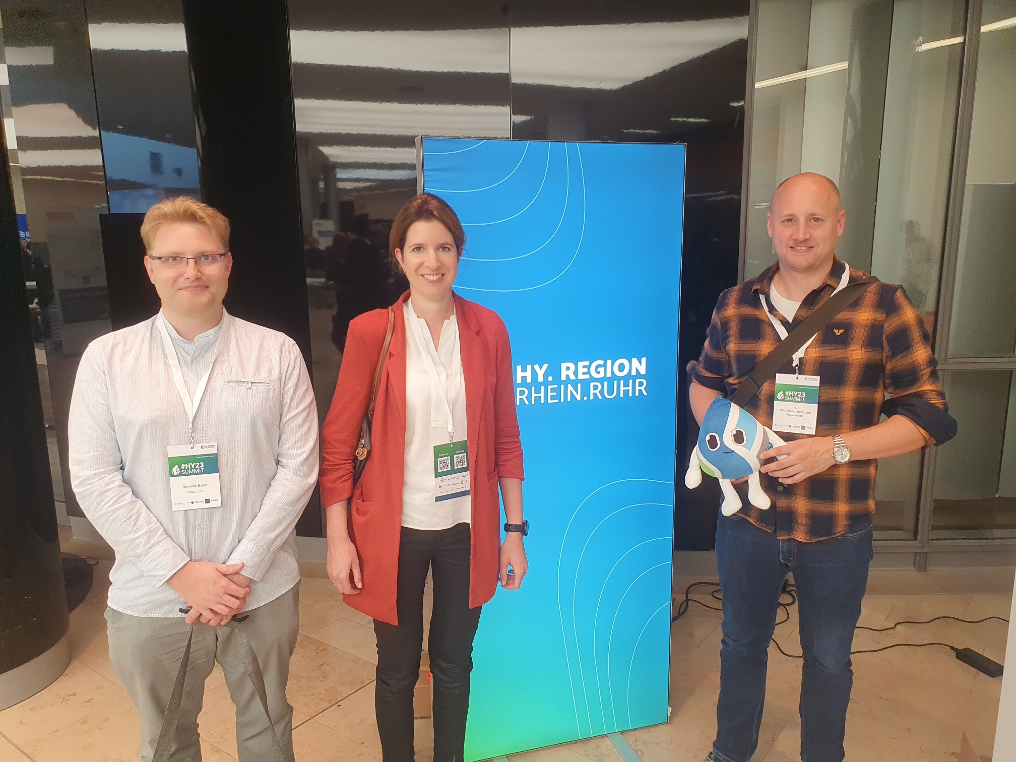 Besuch auf der Hy.Summit.Rhein.Ruhr vom 18.-20. September 2023., 3 Personen mit Aufsteller, wo drauf steht Hy.Summit.Rhein.Ruhrim  im Hintergrund