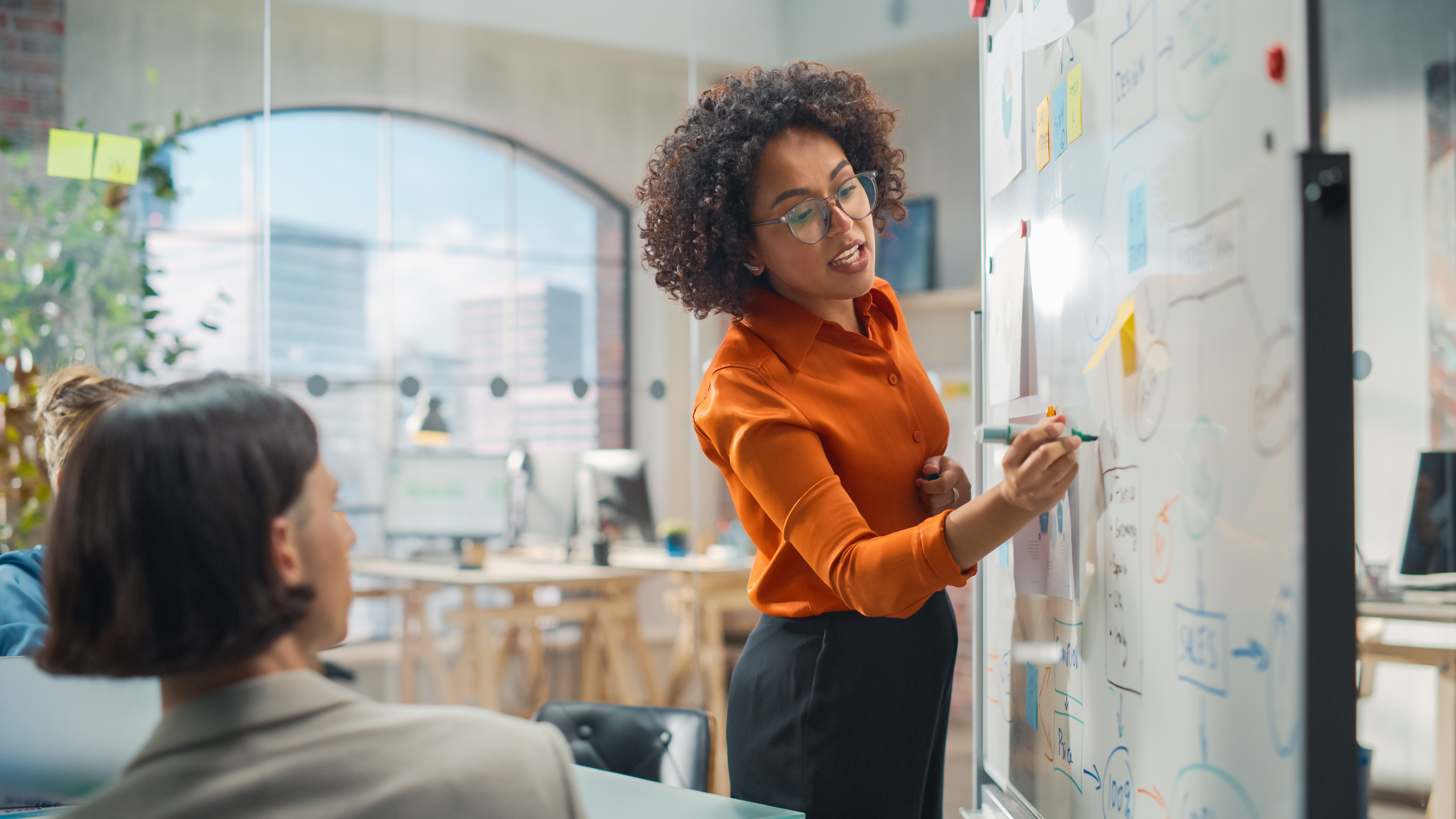 Das Bild zeigt eine Frau vor einem Whiteboard, auf dem Strategien skizziert sind, während sie einer Gruppe etwas erklärt. Sie vermittelt Ideen zur Entwicklung von Open-Source-Strategien, wobei das Whiteboard als visuelles Hilfsmittel für die strukturierte Diskussion und Planung dient.