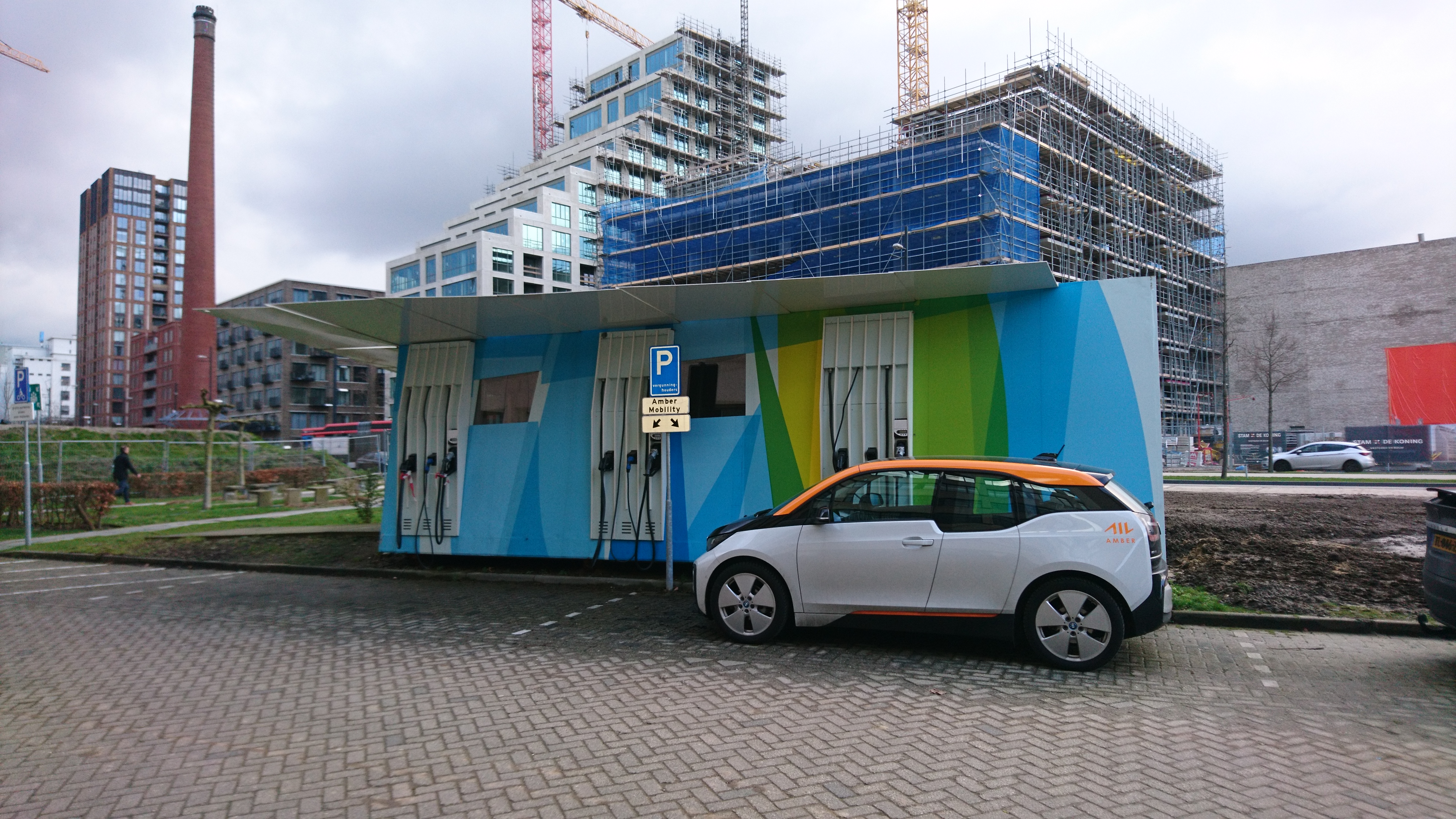Das Bild zeigt ein Elektrofahrzeug an einer großen Ladesäule in einem städtischen Umfeld, umgeben von Baustellen und Gebäuden.  Viele Kommunen fördern Wasserstoff- und Elektromobilität, um umweltfreundliche Fahrzeugangebote zu schaffen und als Vorbild zu fungieren. Der Markt ist dynamisch mit ständigen Entwicklungen. Die kommunale Flotte umfasst Verwaltung und Eigenbetriebe, benötigt spezialisierte Fahrzeuge. Das Fraunhofer IML bietet Unterstützung bei Systemplanung, Flottenintegration, Marktanalysen und der Akquise von Fördermitteln..