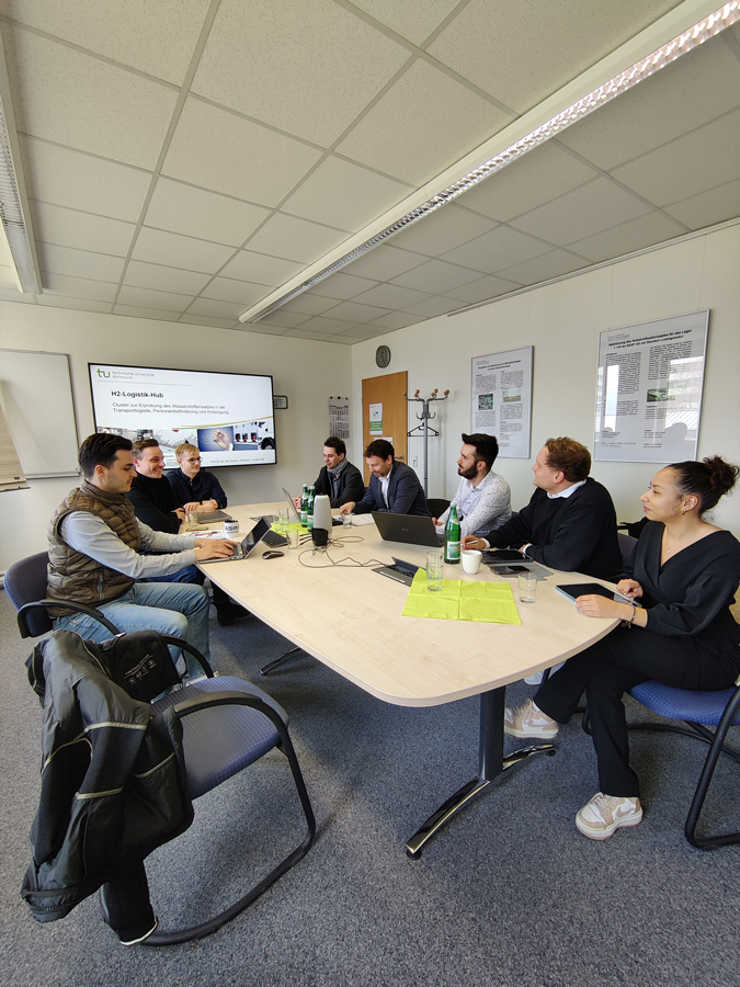 Eine Gruppe von acht Personen sitzt in einem Konferenzraum an einem ovalen Tisch. Auf dem Tisch stehen Laptops, Notizen und Wasserflaschen. Im Hintergrund läuft eine Präsentation zum »H2-Logistik Hub« auf einem Bildschirm.