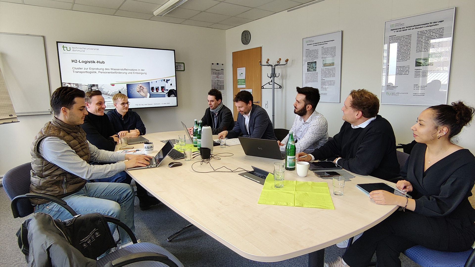 Eine Gruppe von acht Personen sitzt in einem Konferenzraum an einem ovalen Tisch. Auf dem Tisch stehen Laptops, Notizen und Wasserflaschen. Im Hintergrund läuft eine Präsentation zum »H2-Logistik Hub« auf einem Bildschirm.