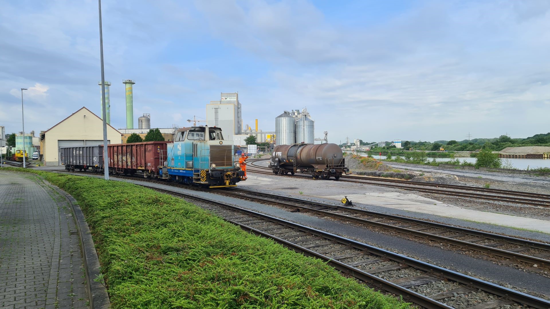 Eine Güterlokomotive, welche zwei Wagen durch ein Hafengebiet zieht. Es sind Gleise und ein weiterer Wagen zu sehen.