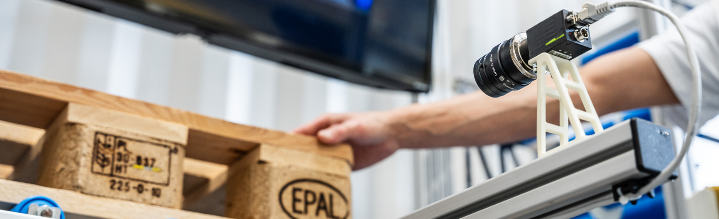 Eine Computer Vision Kamera erfasst eine Holzpalette mit EPAL-Stempel, während ein Bildschirm im Hintergrund Informationen anzeigt