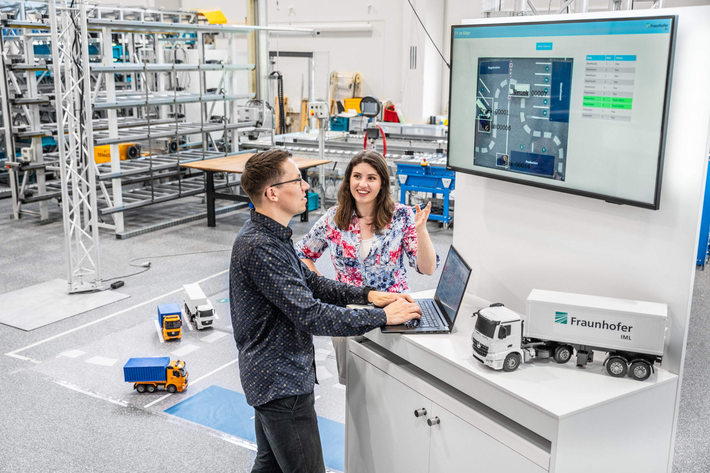 Zwei Forschende, die an einem Laptop arbeiten, während sie eine digitale Anzeige mit einem Steuermodul und Modellen von Lastwagen im Hintergrund betrachten. Hierbei handelt es sich um das Projekt »Yard Lense on Edge«.