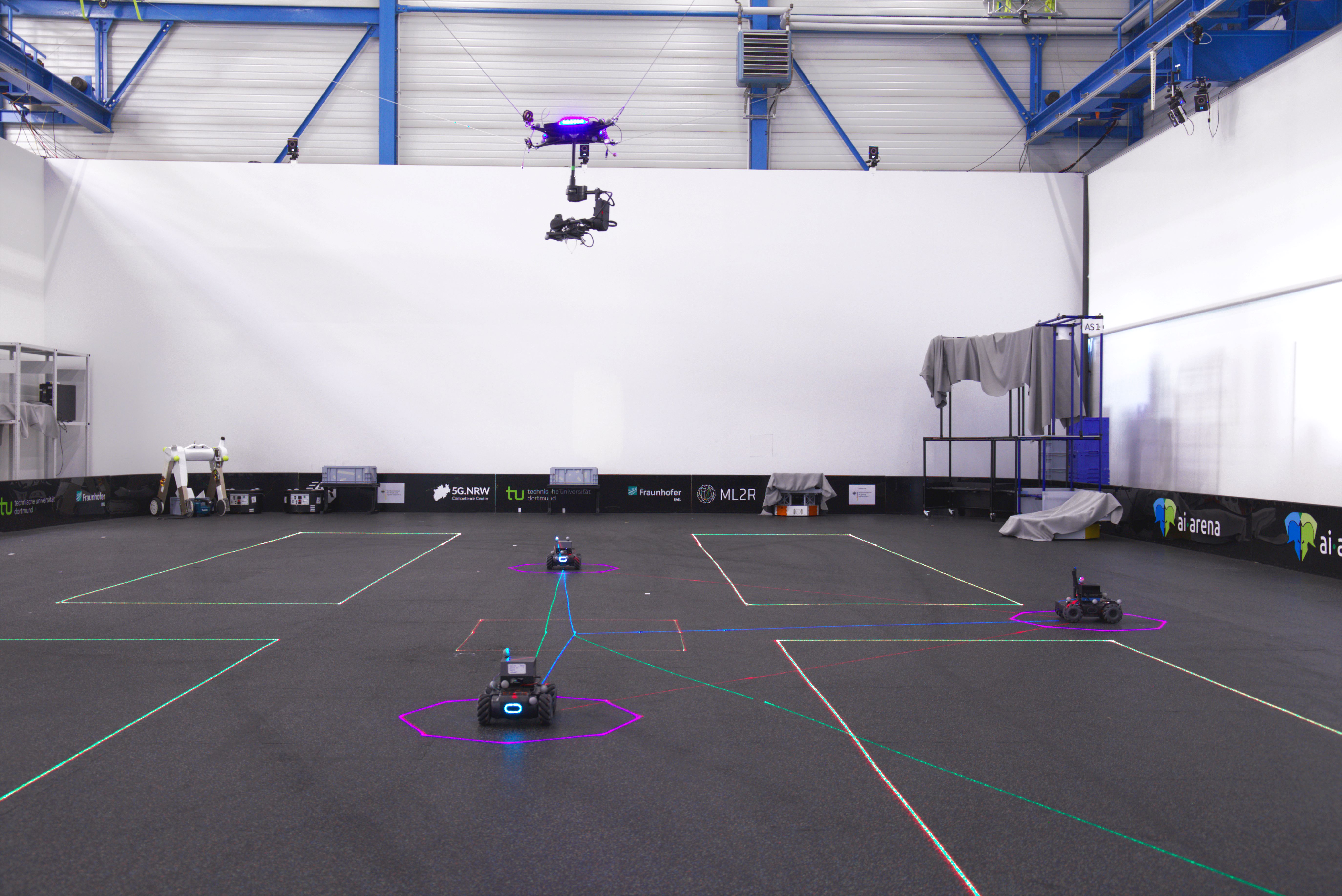 Auf dem Testfeld befinden sich Roboterplattformen, die von Lasern umgeben sind. Darüber schwebt eine Drohne.
