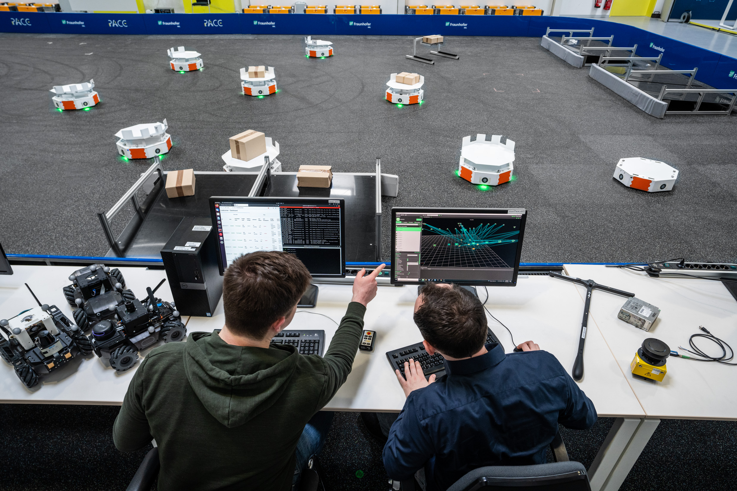 Zwei Menschen von hinten, die auf Bildschirme gucken -davor sind Roboter am Boden