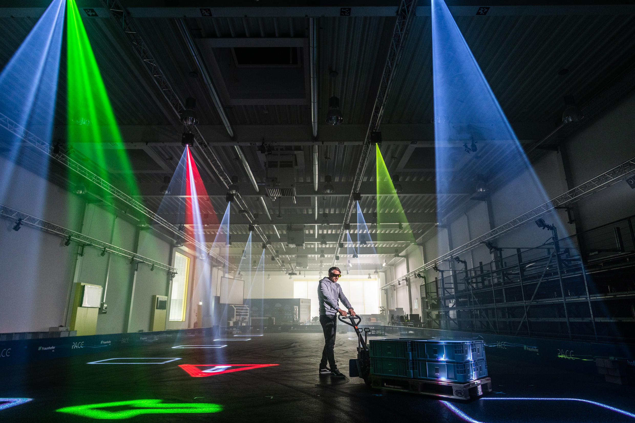 Das Bild zeigt einen Mann, der Paletten durch einen Raum bewegt, wobei ihm Lasermarkierungen am Boden den Weg anzeigen. 