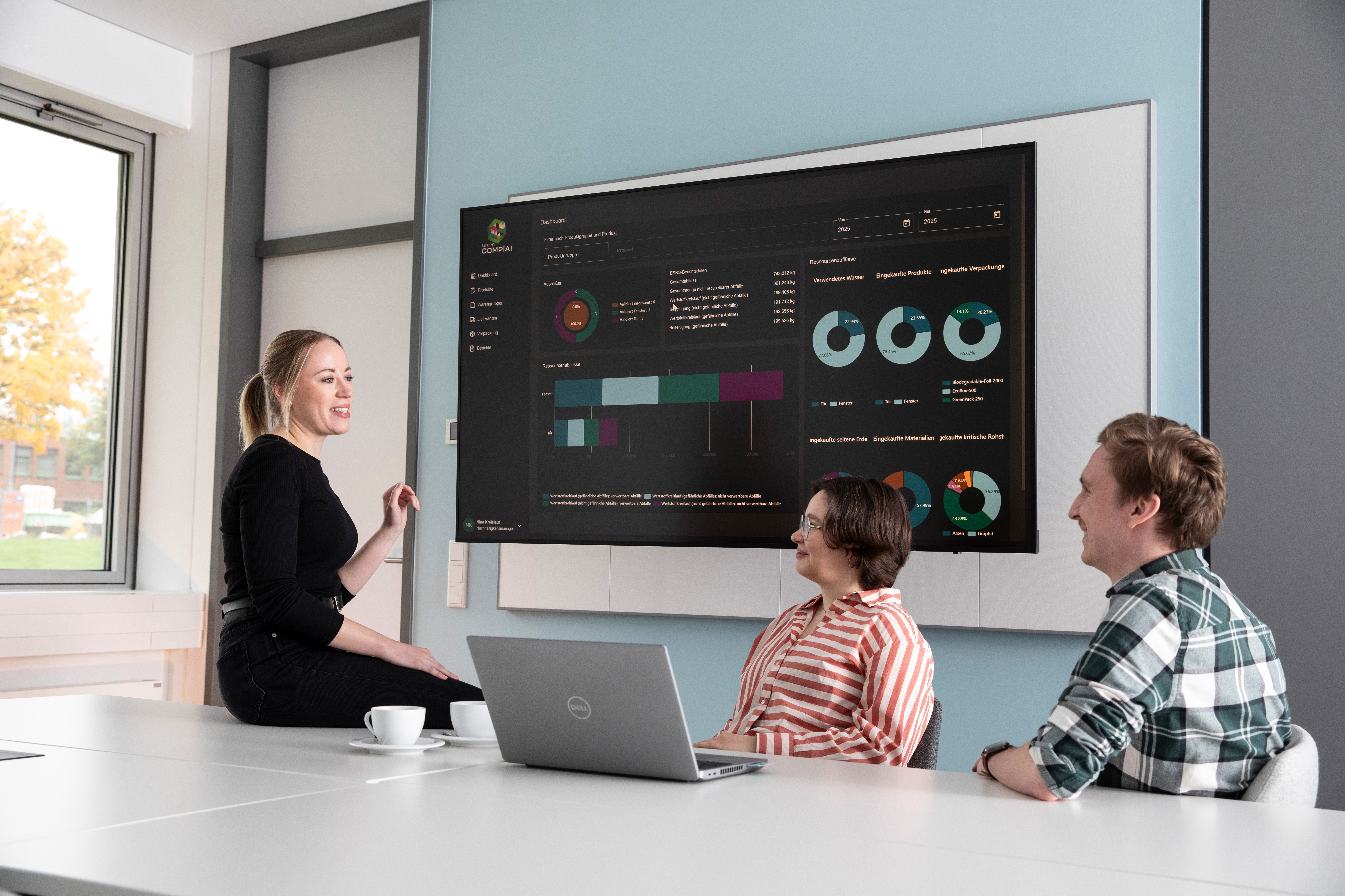 Nachweisführung in Wertschöpfungsnetzwerken Frau lehnt an Tisch und erklärt Frau und Mann Open Source Anwendung an Bildschirm, Nachweisführung in Lieferketten, Analysen und Diagramme, Laptop steht auf Tisch, Menschen lächeln