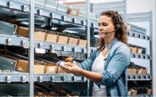 Teaser » Effizienzsteigerung in der Logistik« Eine Frau im Lager trägt ein Headset und nutzt einen Handscanner, um Kartons vor voll bestückten Regalen zu überprüfen.