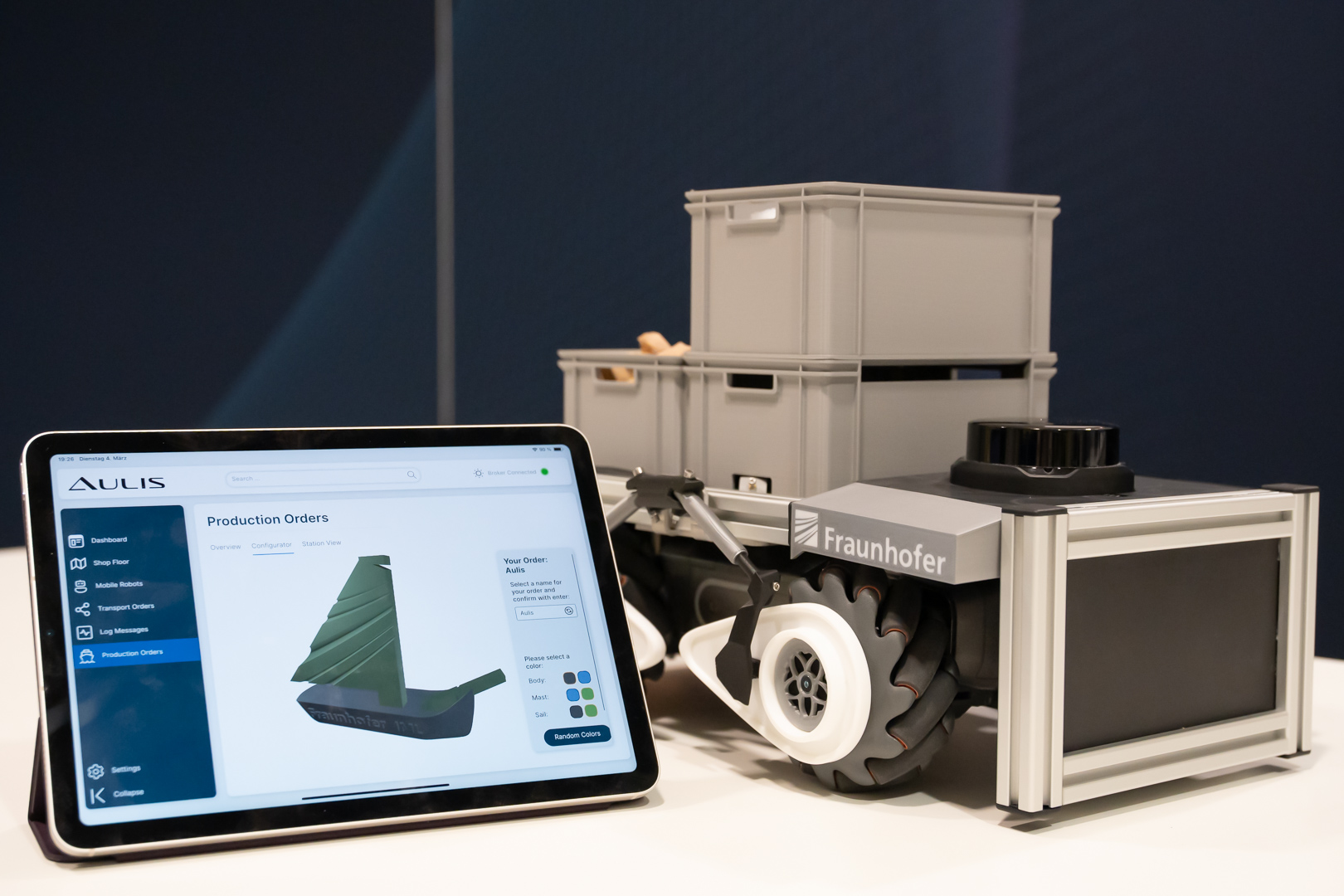 Auf dem Bild ist das Lieitexponat für die LogiMAT des Fraunhofer IML zu sehen: Hände mit einem Tablett zu sehen, auf dem das Programm AULIS läuftt, mit einem Roboter im Hintergrund, auf dem sich Kisten befinden. 