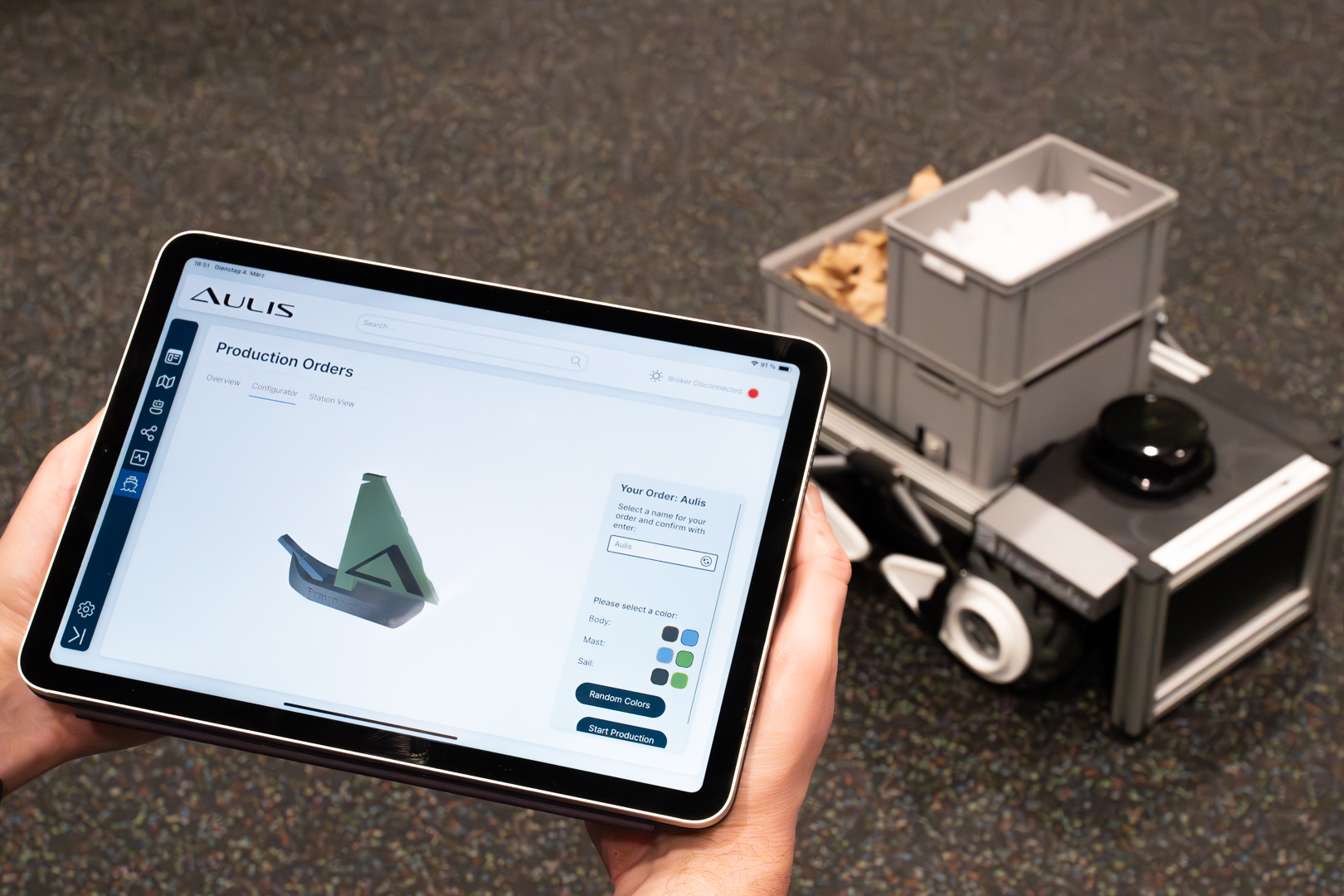 Auf dem Bild ist das Lieitexponat für die LogiMAT des Fraunhofer IML zu sehen: Hände mit einem Tablett zu sehen, auf dem das Programm AULIS läuftt, mit einem Roboter im Hintergrund, auf dem sich Kisten befinden. 
