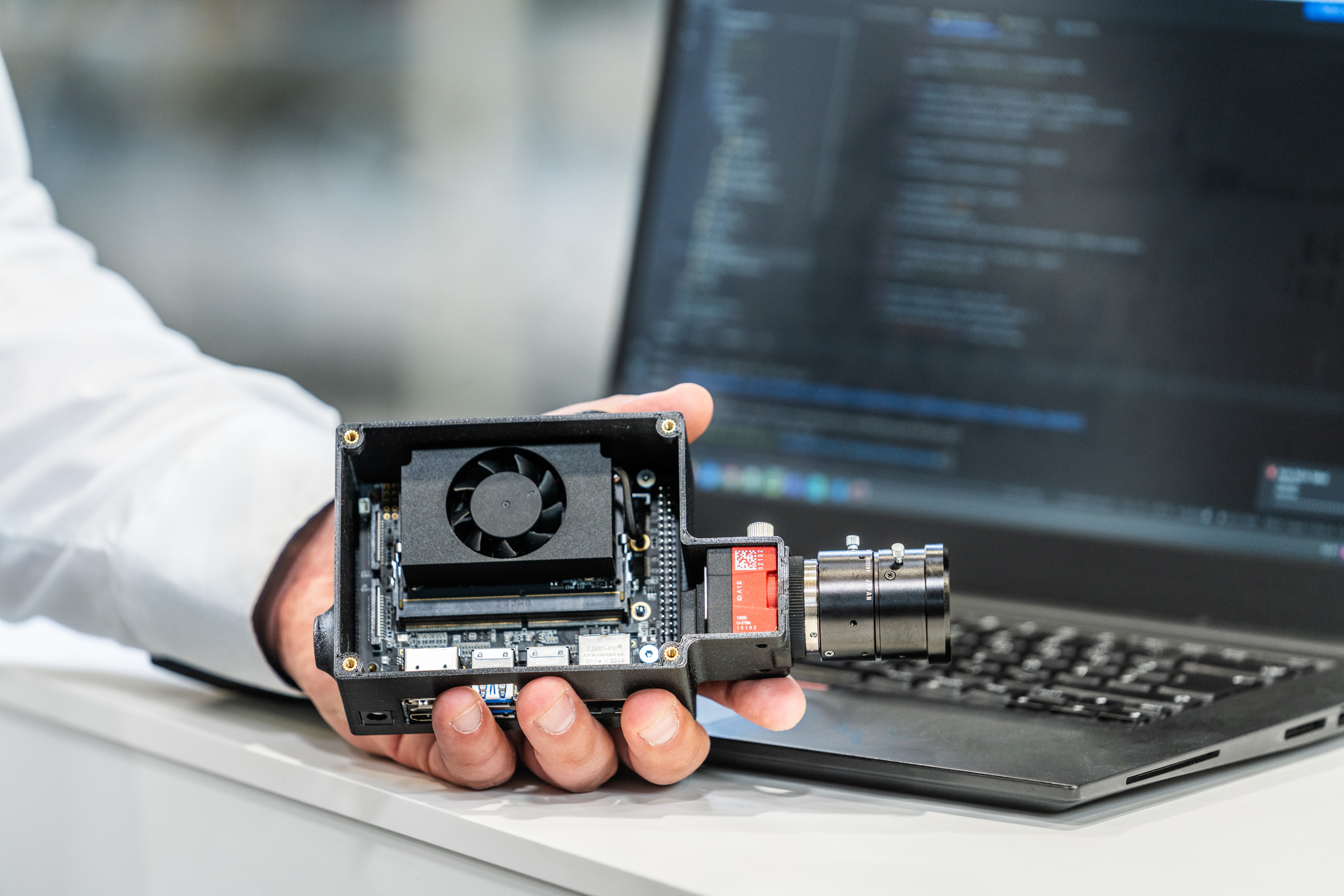 A person is holding a computer vision camera in their hand, while a laptop with programming code can be seen in the background.