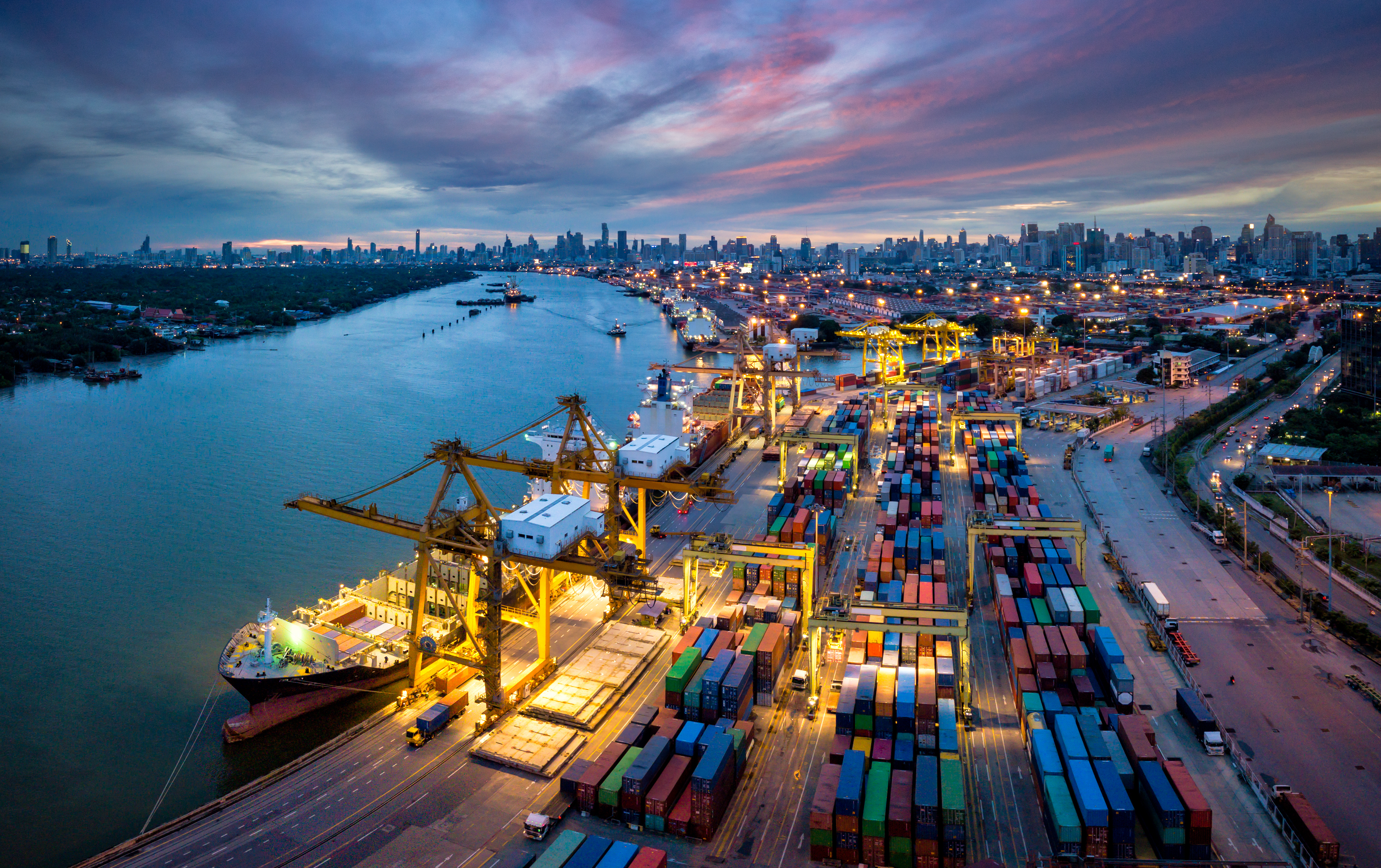 Das Bild zeigt einen großen Frachthafen bei Sonnenuntergang, mit Containerschiffen und gestapelten Containern entlang des Kais, während die Lichter der Stadt im Hintergrund leuchten.