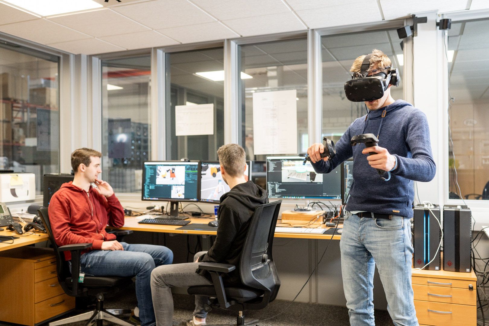 Two men are looking at computer screens, while another man is using VR glasses and controlers.