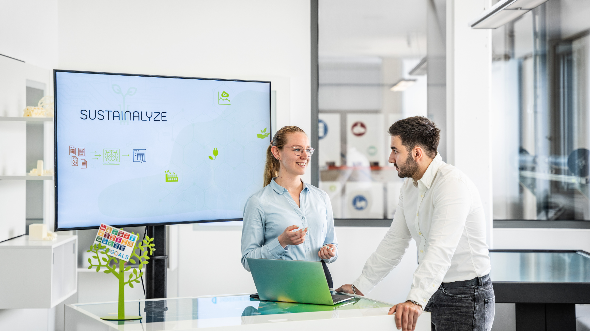 Two people in a modern office standing at a table and talking, with a screen displaying the word ‘SUSTAINABILITY’ visible in the background.
