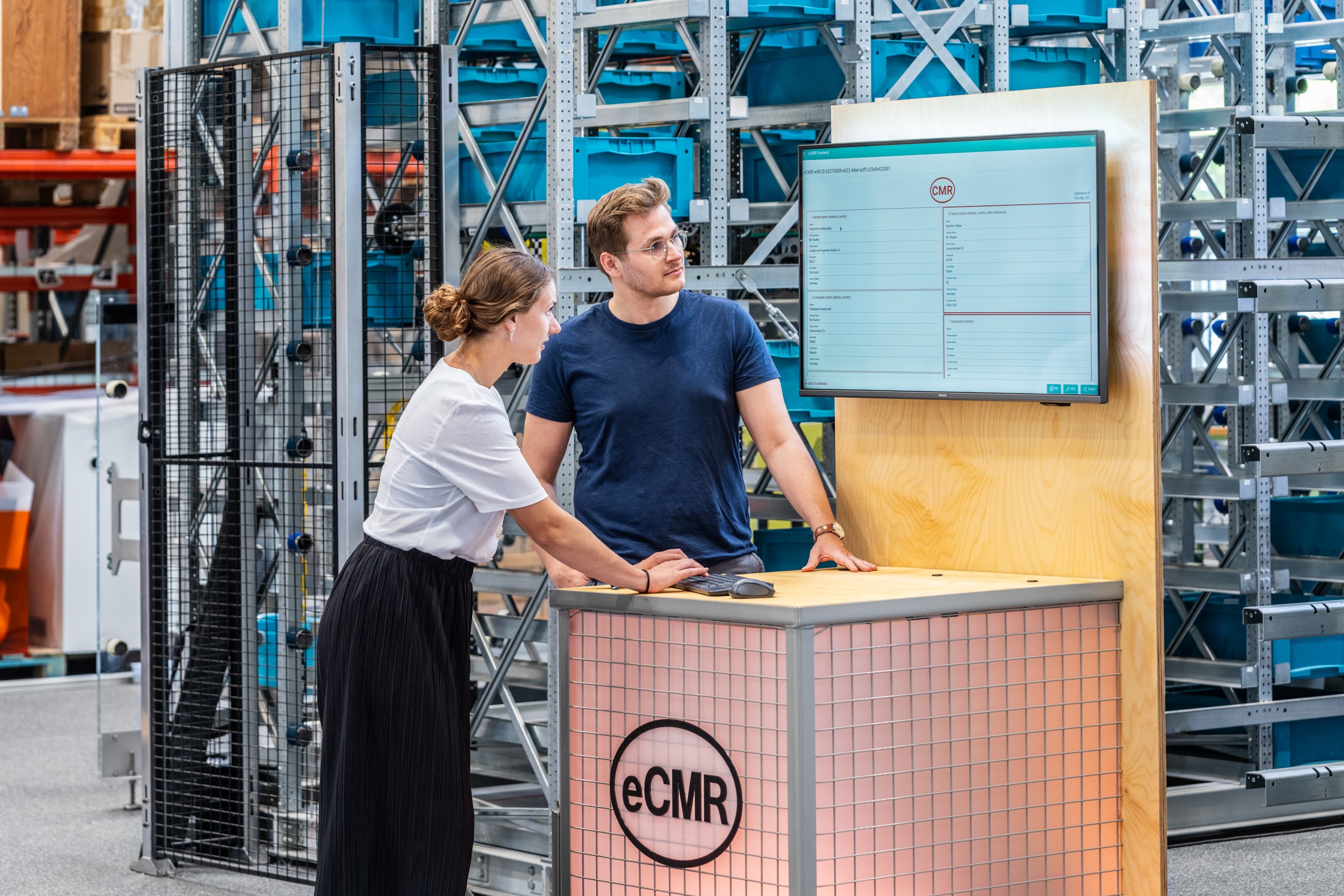 A woman and a man stand at an exhibit with a screen in the background while talking about the eCMR system.