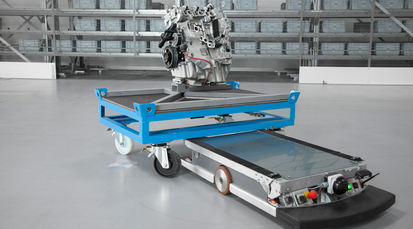 The picture shows a mobile transport device carrying an engine assembly on a blue platform. The platform is mounted on a mobile undercarriage equipped with large wheels. In the background, shelves with several storage containers can be seen, indicating an industrial environment. The entire arrangement appears to be designed for the transport of components within a production hall.